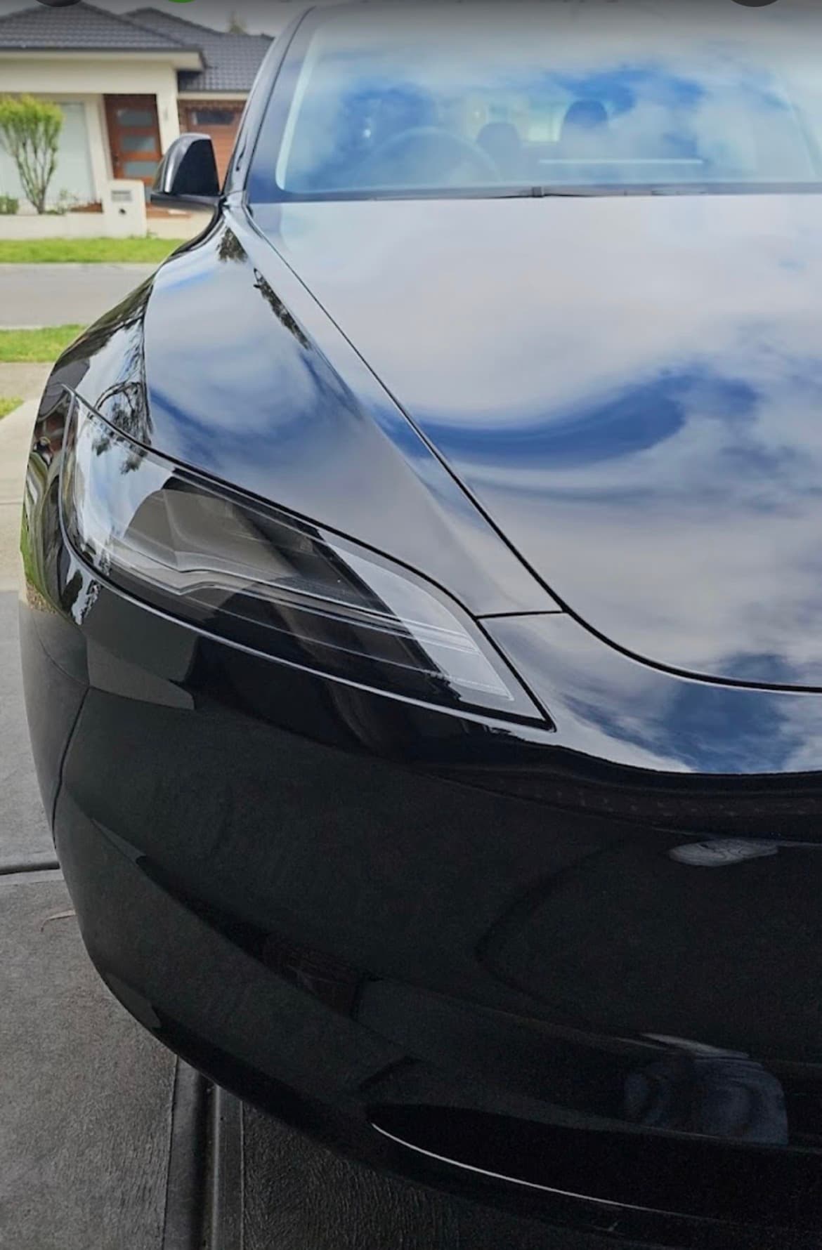 Close-up of a glossy black car's headlight reflecting a blue sky with white clouds.