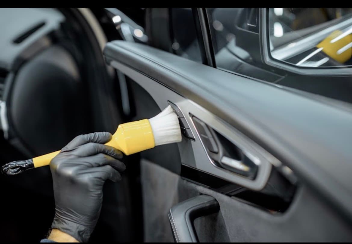 Gloved hand cleaning an interior car door panel with a yellow detailing brush.