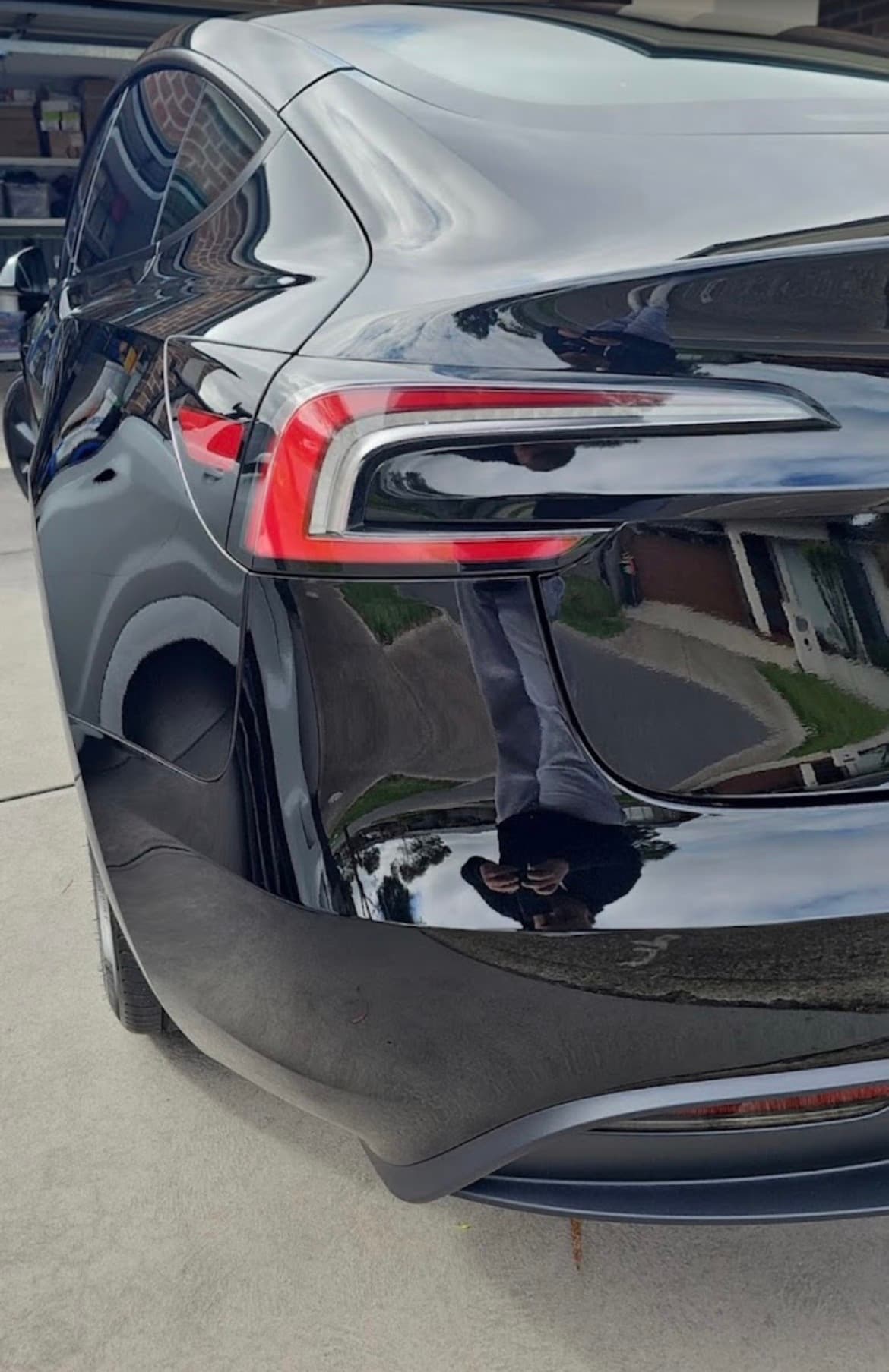 Rear taillight of a glossy black Tesla reflecting a person and a suburban neighborhood.